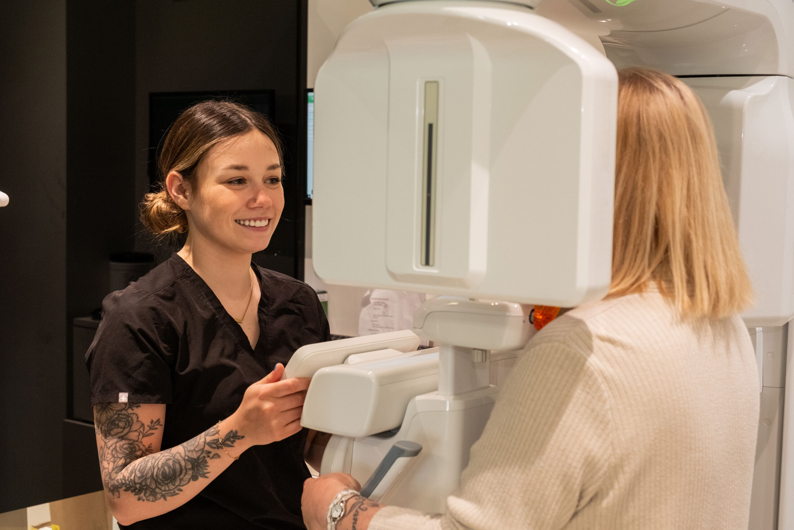 'http://A%20smiling%20healthcare%20professional%20in%20black%20scrubs%20assists%20a%20patient%20with%20a%20large%20medical%20imaging%20machine%20in%20a%20clinical%20setting.%20The%20patient%20faces%20the%20machine,%20while%20the%20professional%20provides%20guidance.