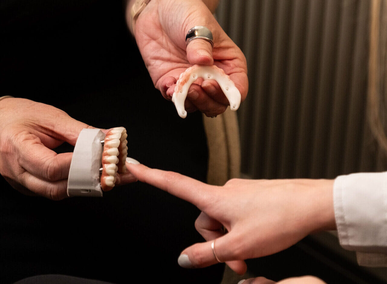 'http://Two%20hands%20holding%20dental%20prosthetics,%20one%20with%20an%20upper%20denture%20and%20the%20other%20with%20a%20lower%20denture,%20while%20a%20third%20hand%20points%20toward%20the%20models.