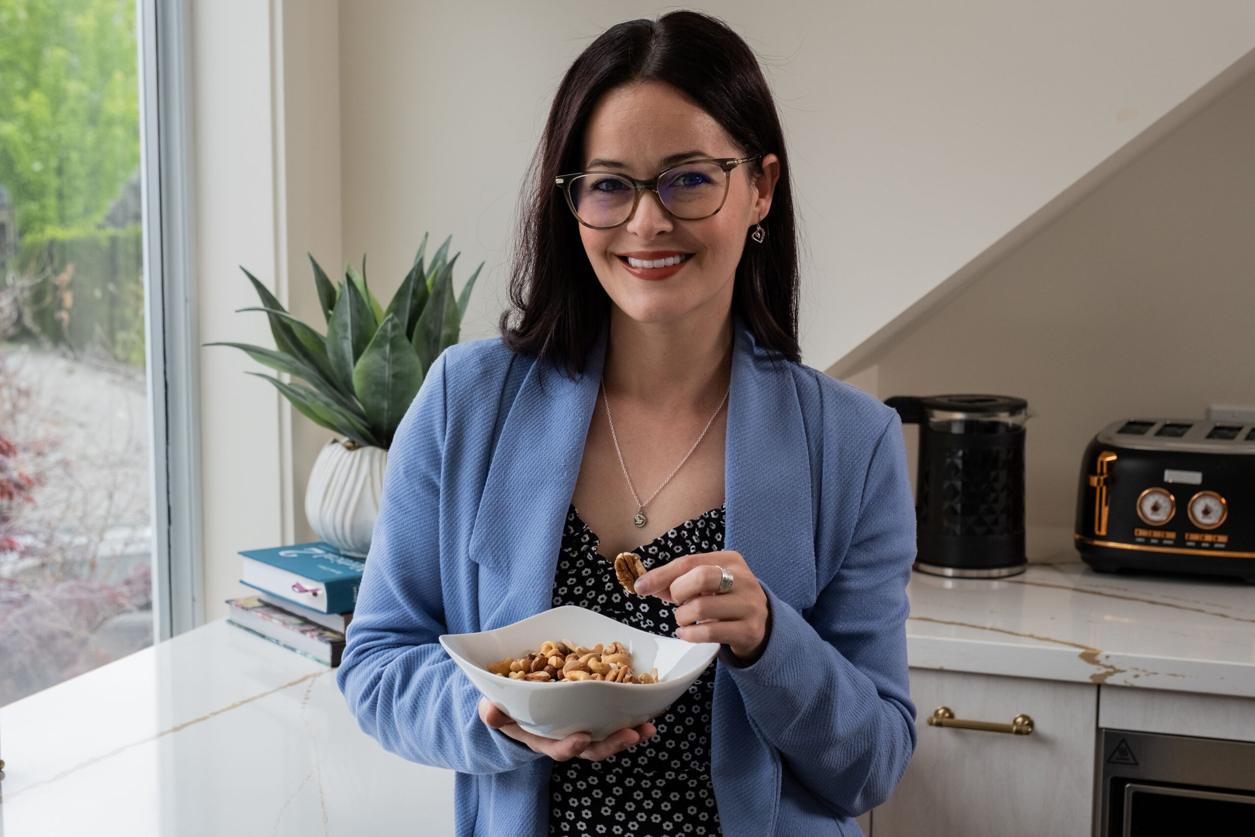 'http://A%20woman%20with%20dark%20hair%20and%20glasses,%20wearing%20a%20blue%20blazer%20and%20polka-dot%20top,%20smiles%20while%20holding%20a%20bowl%20of%20mixed%20nuts.%20A%20potted%20plant,%20books,%20and%20a%20coffee%20maker%20are%20in%20the%20background.