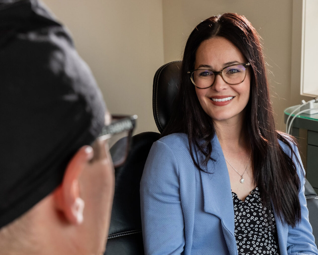 'http://A%20woman%20with%20long%20dark%20hair,%20glasses,%20and%20a%20blue%20blazer%20smiles%20while%20sitting%20across%20from%20another%20person%20in%20a%20casual%20indoor%20setting,%20suggesting%20a%20friendly%20conversation%20or%20counseling%20session.