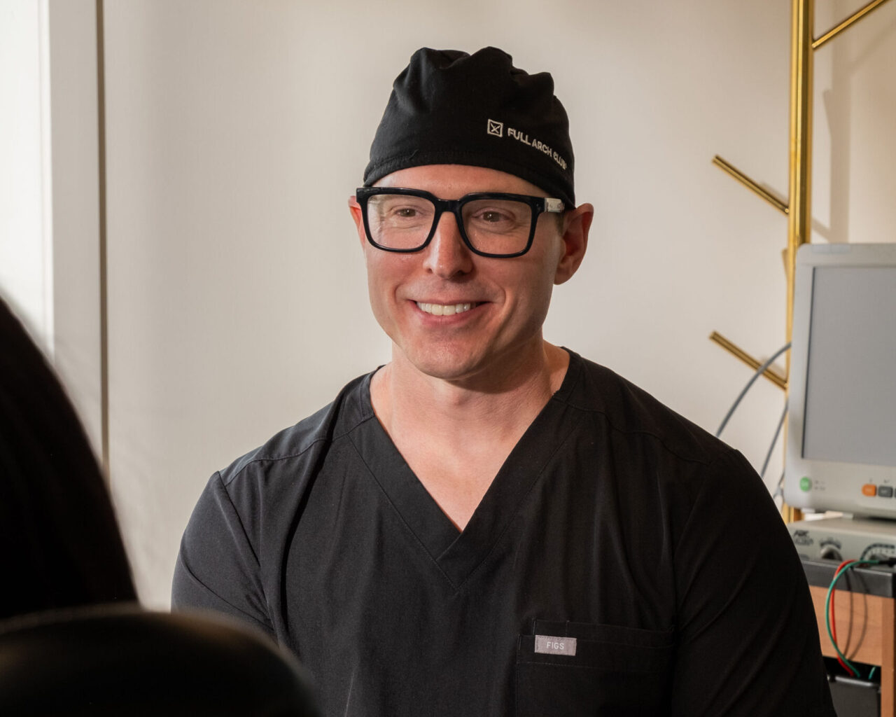 'http://A%20person%20wearing%20black%20medical%20scrubs,%20a%20black%20surgical%20cap,%20and%20glasses%20smiles%20while%20sitting%20indoors,%20with%20medical%20equipment%20visible%20in%20the%20background.