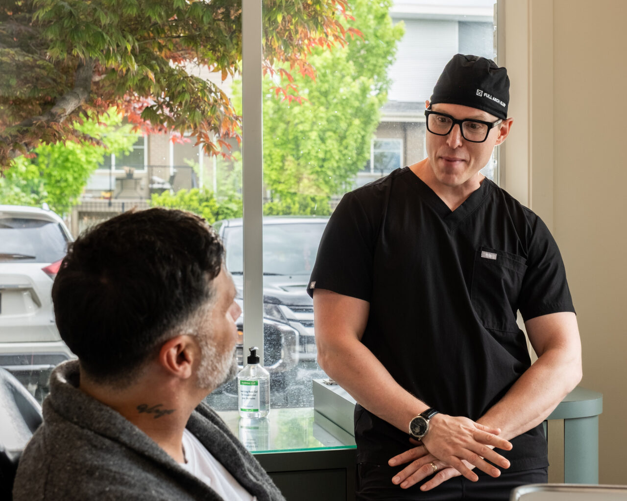 'http://A%20man%20in%20black%20medical%20scrubs%20and%20glasses%20speaks%20to%20a%20seated%20man%20with%20a%20neck%20tattoo%20in%20a%20bright%20office%20with%20large%20windows%20and%20trees%20visible%20outside.