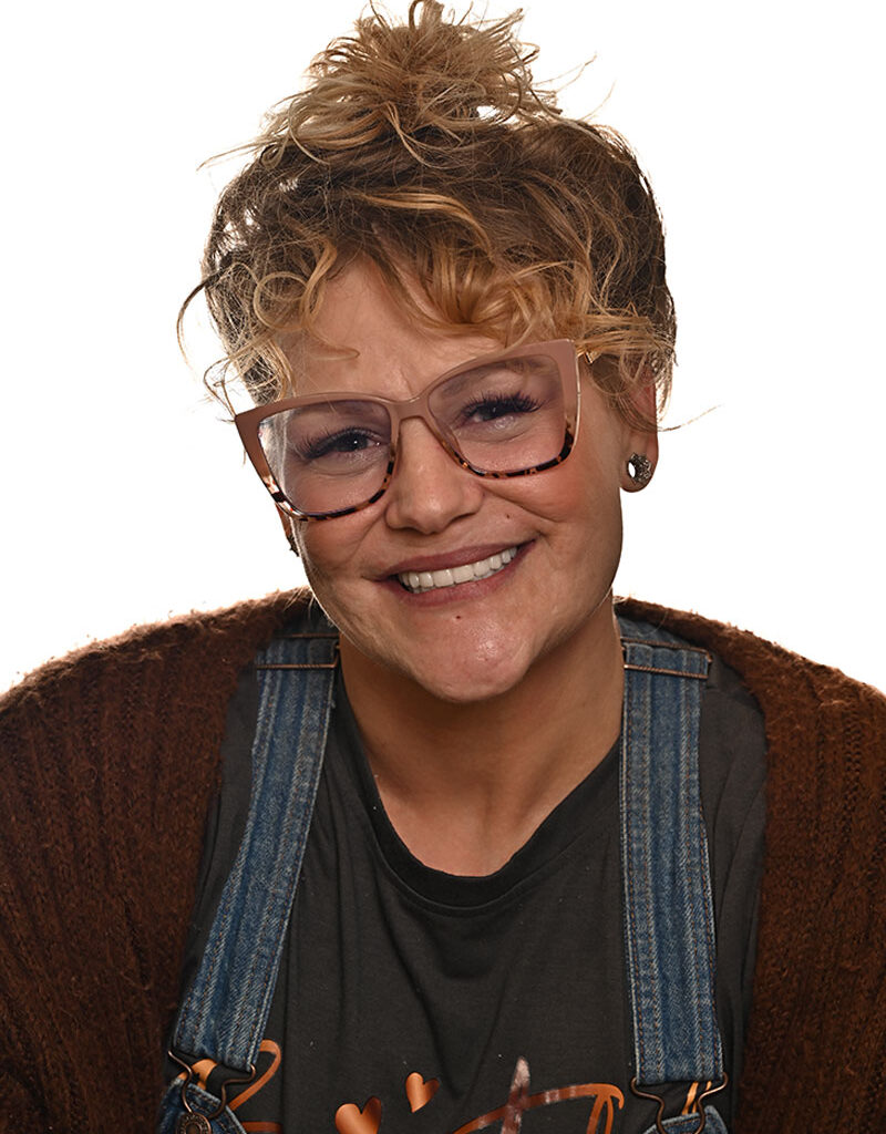 A smiling person with curly blonde hair in a messy bun, wearing oversized clear-framed glasses, denim overalls, a brown sweater, and a black t-shirt, posing against a white background.