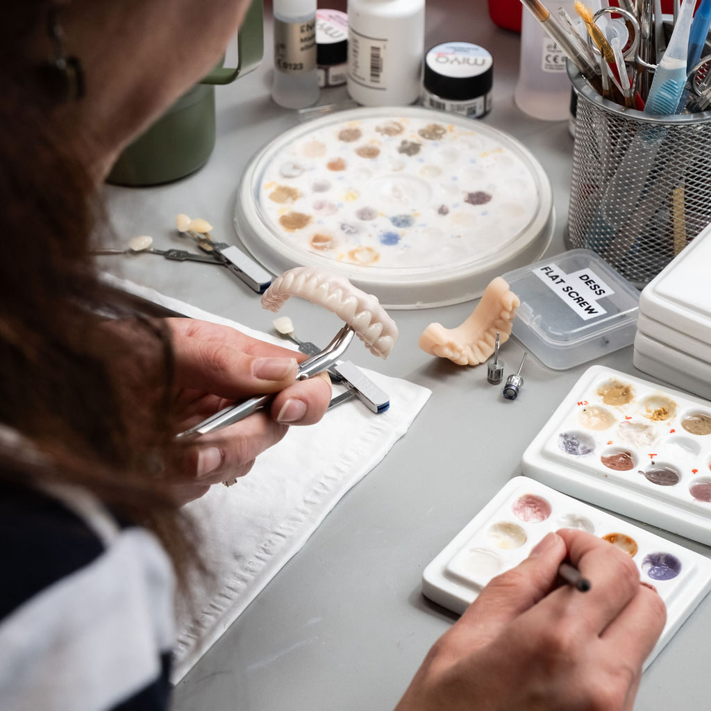 A person works at a table with dental tools, artificial teeth, and palettes of paint, carefully painting a dental prosthetic.
