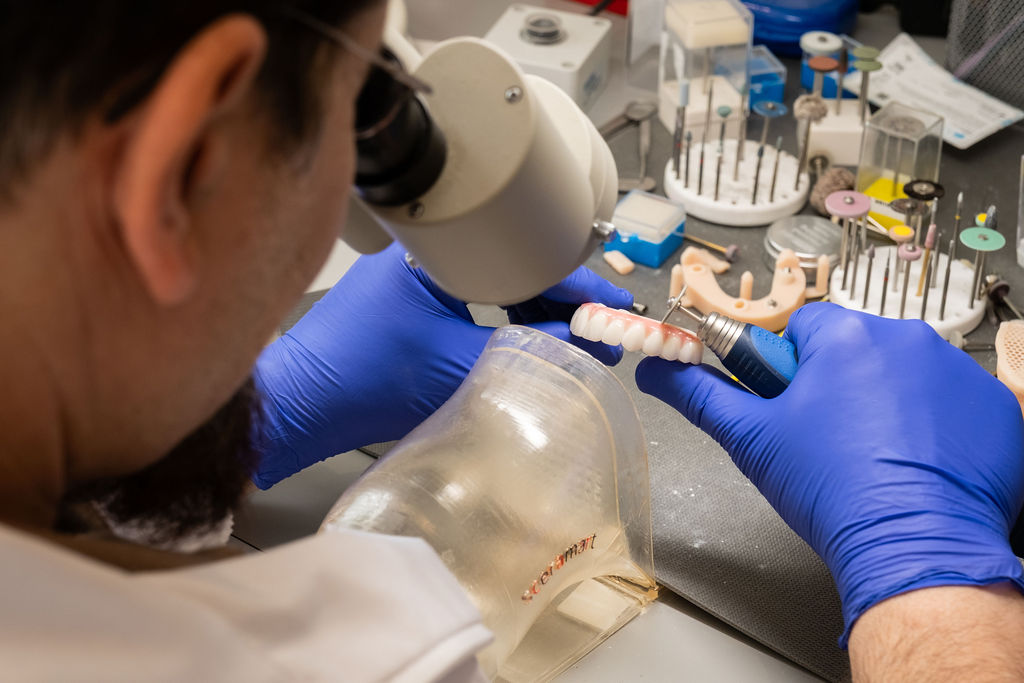A dental technician wearing blue gloves uses a tool to work on a set of artificial teeth, with various dental instruments and equipment visible on the workbench.