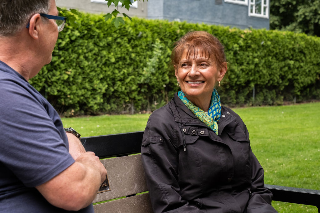 A woman with short brown hair and a colorful scarf smiles while sitting on a park bench, with green grass, a hedge, and a building in the background.