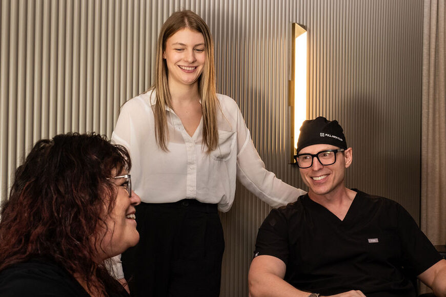 'http://Three%20people%20are%20having%20a%20friendly%20conversation%20indoors.%20A%20woman%20in%20a%20white%20blouse%20stands%20smiling%20between%20a%20seated%20woman%20with%20glasses%20and%20a%20man%20in%20black%20scrubs%20and%20a%20surgical%20cap.%20The%20atmosphere%20is%20warm%20and%20professional.