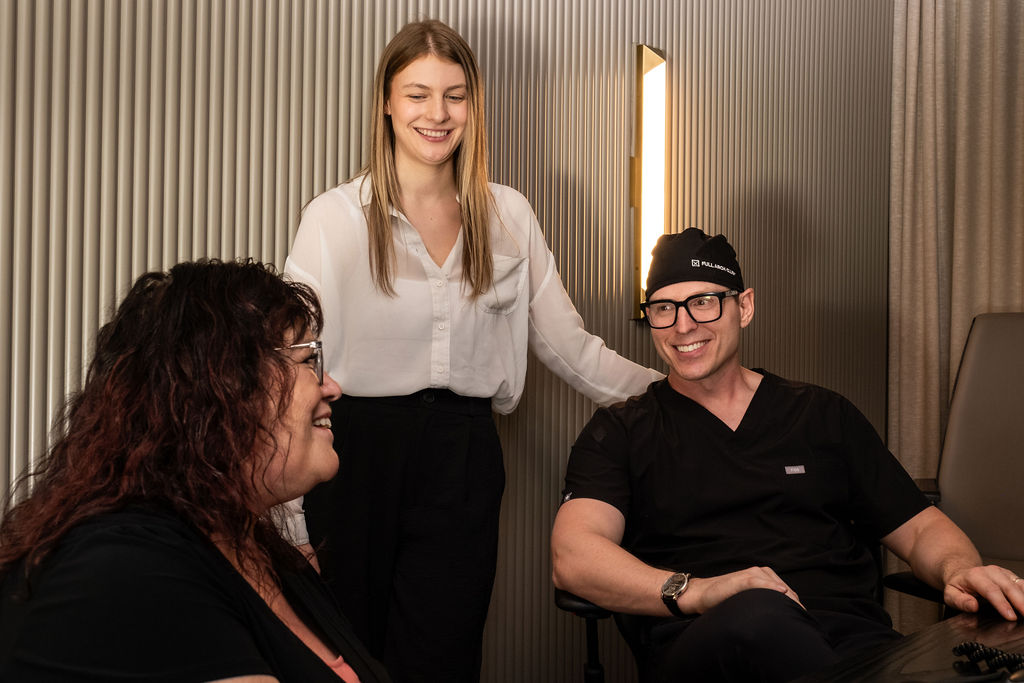 'http://Three%20people%20are%20in%20a%20modern%20office%20setting,%20smiling%20and%20talking.%20One%20woman%20is%20seated,%20another%20woman%20stands%20beside%20her,%20and%20a%20man%20in%20black%20scrubs%20and%20glasses%20sits%20at%20a%20desk,%20all%20appearing%20engaged%20and%20friendly.