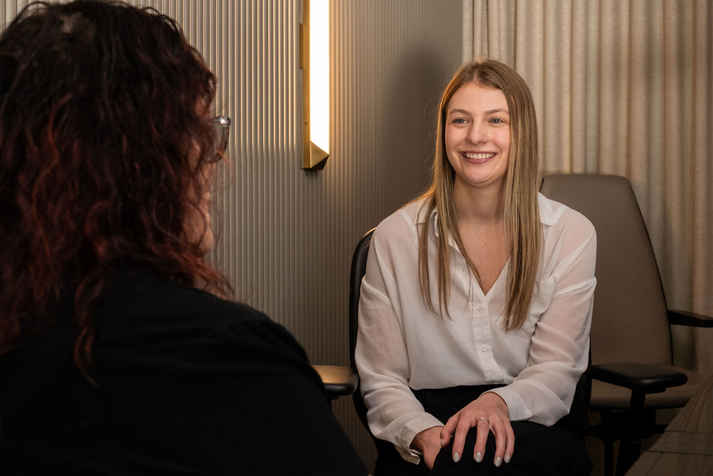 'http://Two%20women%20are%20sitting%20in%20an%20office,%20having%20a%20conversation.%20One%20woman,%20facing%20the%20camera,%20is%20smiling%20and%20wearing%20a%20white%20blouse.%20The%20other%20woman%20is%20partially%20visible%20from%20behind.