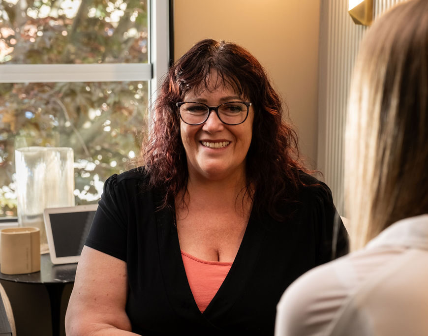 'http://A%20woman%20with%20glasses%20and%20dark,%20wavy%20hair%20smiles%20while%20sitting%20across%20from%20another%20person%20in%20an%20indoor%20setting,%20with%20a%20window%20and%20desk%20visible%20in%20the%20background.