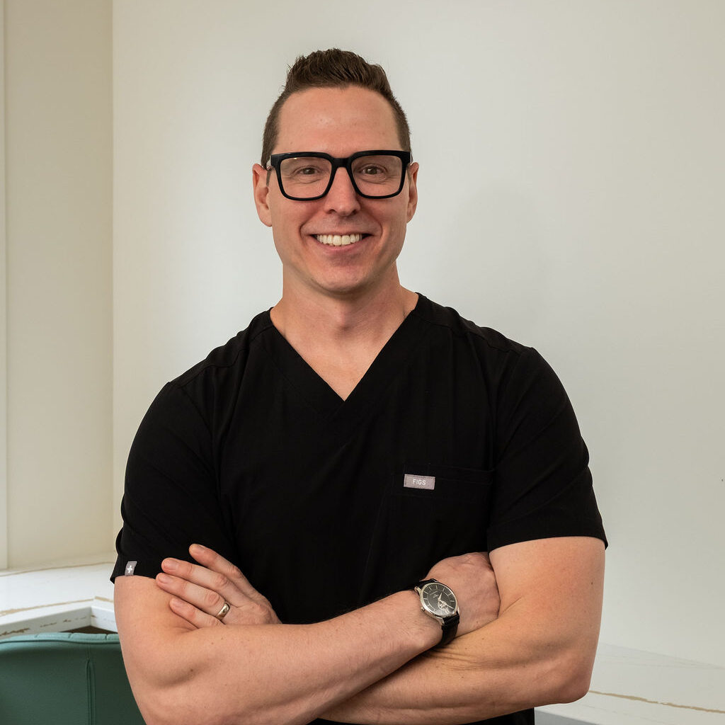 'http://A%20smiling%20man%20wearing%20glasses%20and%20a%20black%20scrub%20top%20stands%20with%20arms%20crossed%20in%20a%20bright,%20modern%20room.