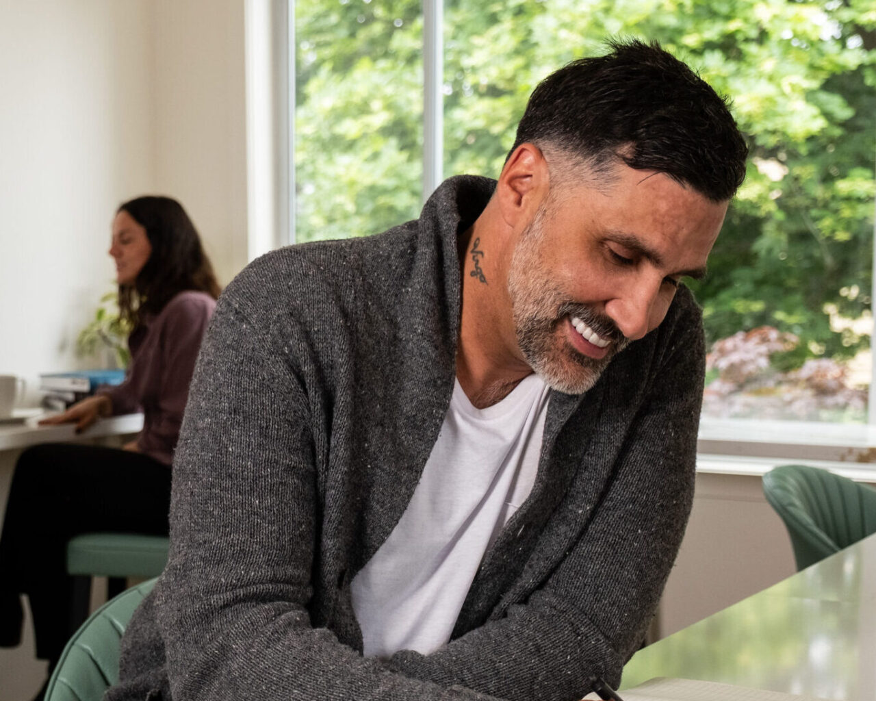 'http://A%20man%20with%20tattoos,%20wearing%20a%20gray%20cardigan%20and%20white%20shirt,%20smiles%20as%20he%20writes%20in%20a%20notebook%20at%20a%20table%20near%20a%20large%20window.%20Another%20person%20sits%20in%20the%20background.%20Green%20trees%20are%20visible%20outside.