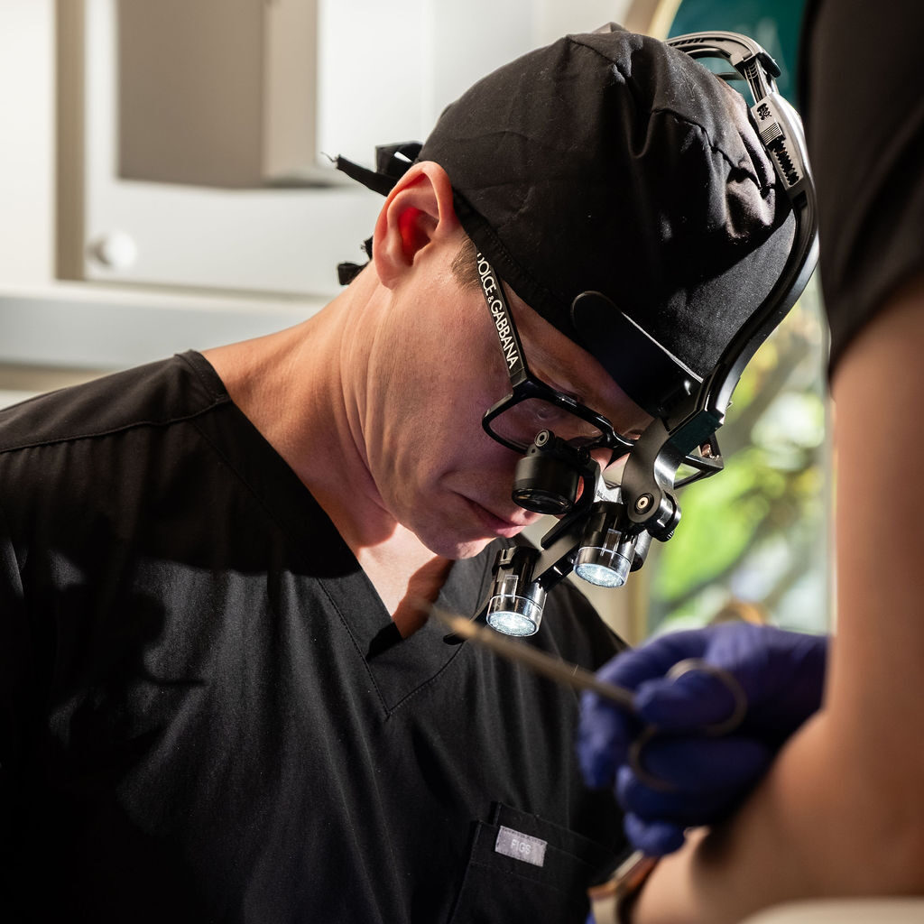 'http://A%20dentist%20wearing%20magnifying%20glasses%20and%20a%20headlamp%20focuses%20intently%20while%20performing%20a%20procedure,%20dressed%20in%20black%20scrubs%20and%20gloves,%20with%20a%20blurred%20patient%20or%20assistant%20in%20the%20foreground.