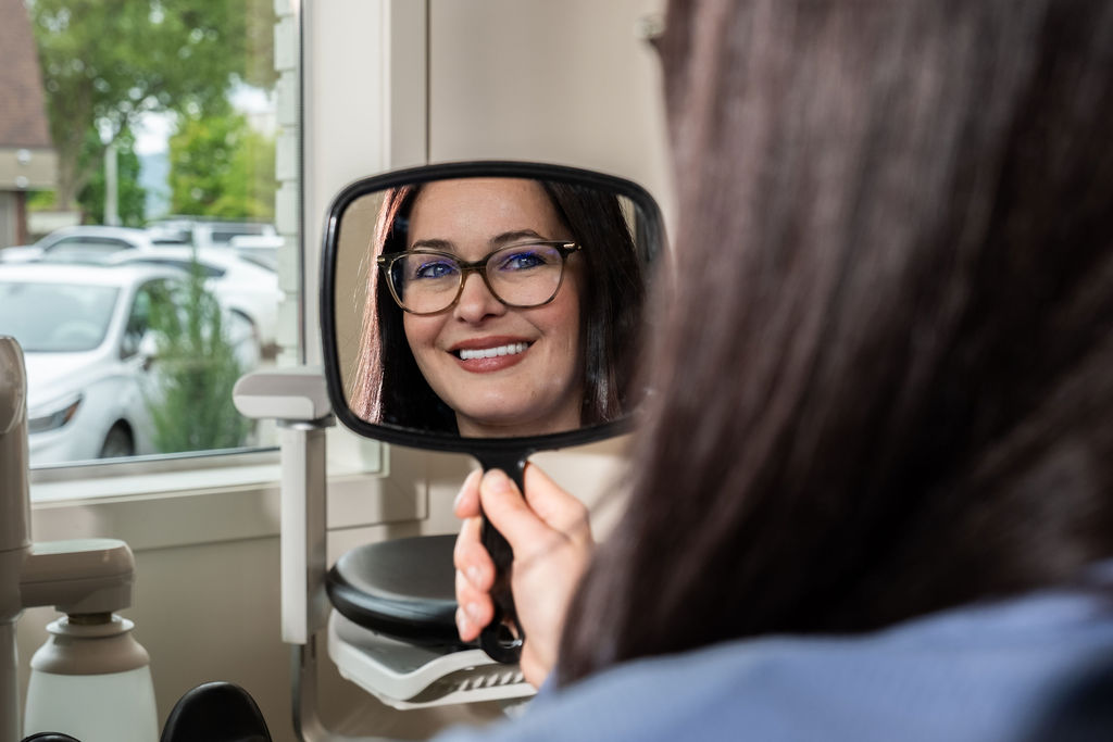 A woman with long brown hair and glasses smiles while looking at her reflection in a handheld mirror at what appears to be an optometrist’s office, with a window and parked cars in the background.