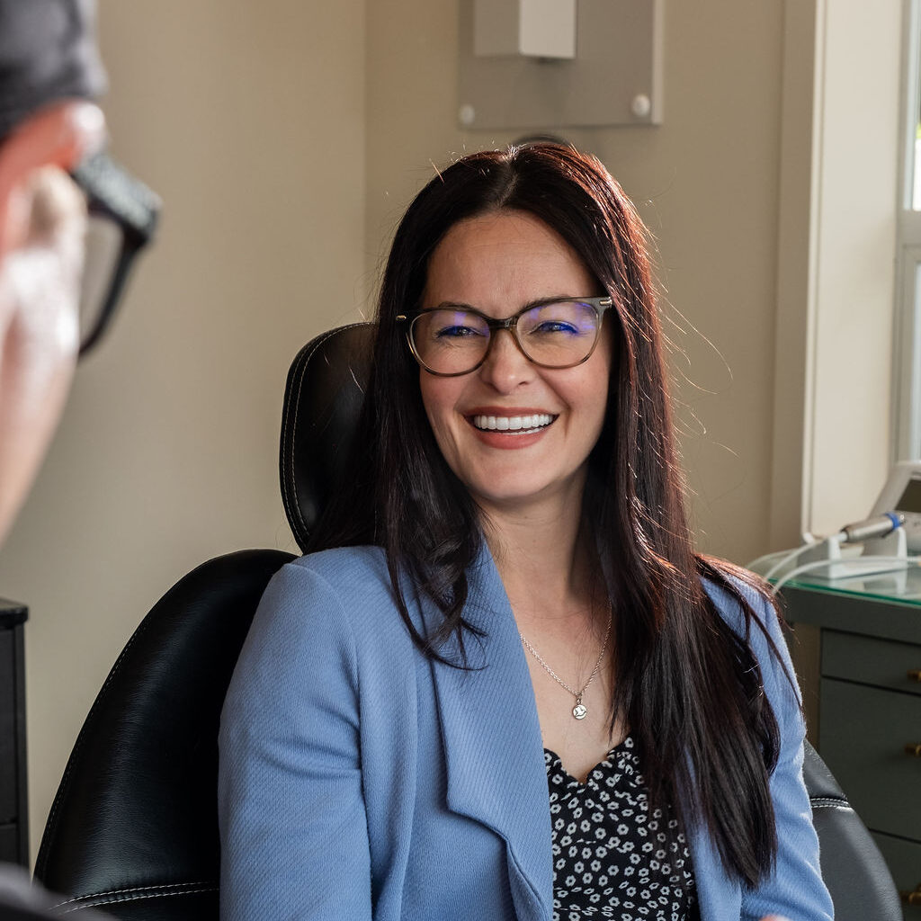 'http://A%20woman%20with%20long%20dark%20hair%20and%20glasses,%20wearing%20a%20blue%20blazer%20and%20patterned%20top,%20smiles%20while%20sitting%20in%20an%20office%20chair,%20engaging%20in%20conversation%20with%20another%20person%20whose%20hands%20are%20visible%20in%20the%20foreground.