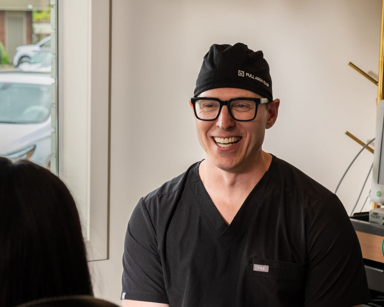 'http://A%20smiling%20man%20wearing%20glasses,%20a%20black%20scrub%20top,%20and%20a%20black%20cap%20sits%20indoors,%20talking%20to%20another%20person%20whose%20back%20is%20to%20the%20camera.%20Natural%20light%20comes%20through%20a%20nearby%20window.