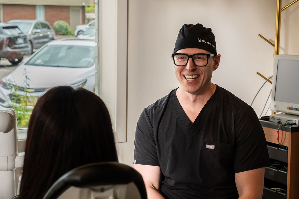 'http://A%20smiling%20man%20in%20black%20scrubs%20and%20a%20cap%20speaks%20with%20a%20woman%20in%20an%20office%20with%20medical%20equipment,%20near%20a%20window%20showing%20parked%20cars%20outside.