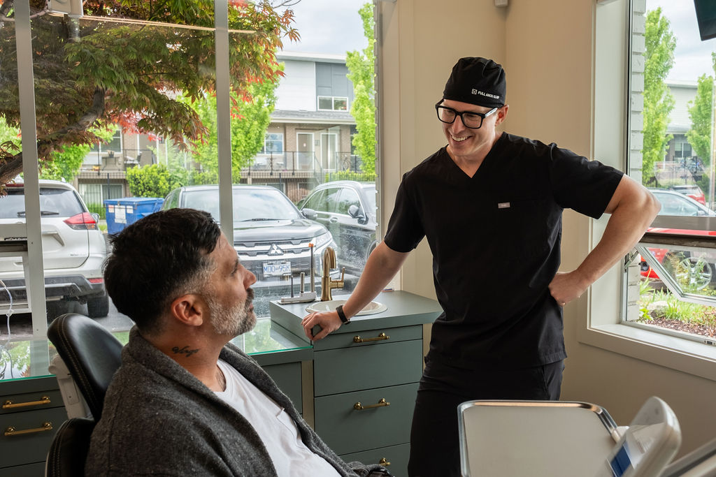 A dentist in black scrubs and glasses smiles while talking to a patient sitting in a dental chair near a large window that shows parked cars and trees outside.