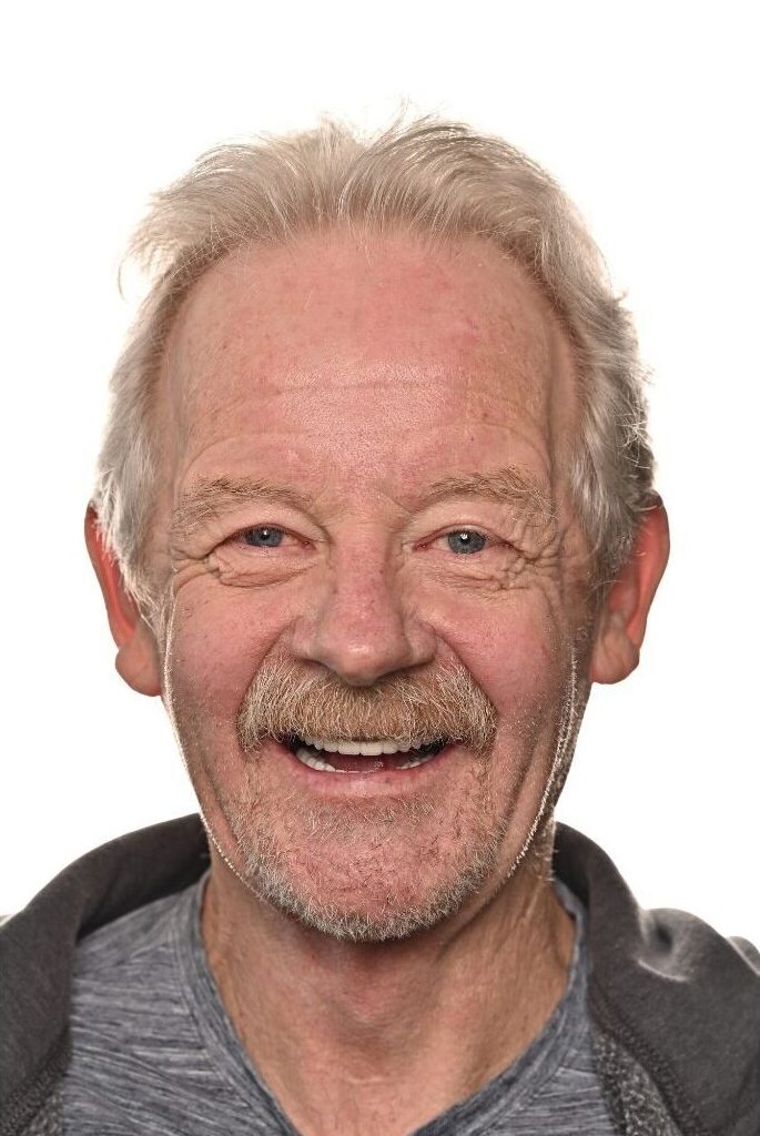 An older man with light hair and a mustache smiles warmly at the camera against a plain white background. He is wearing a gray shirt and dark jacket.