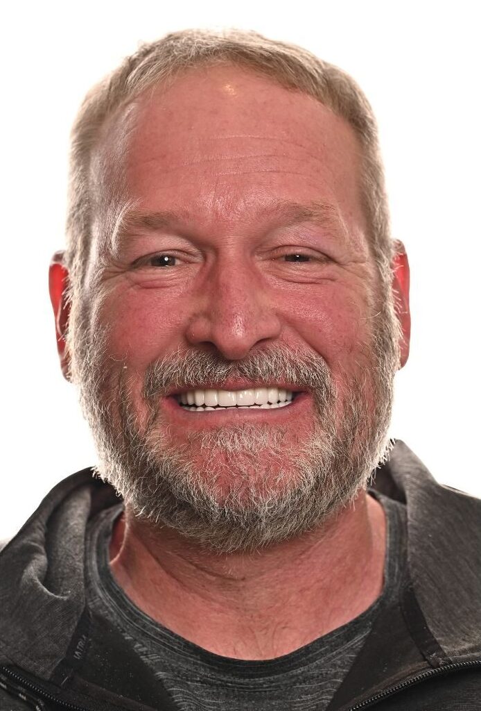 A middle-aged man with short, light hair and a gray beard is smiling broadly, showing his teeth. He is wearing a dark hoodie and is photographed against a plain white background.