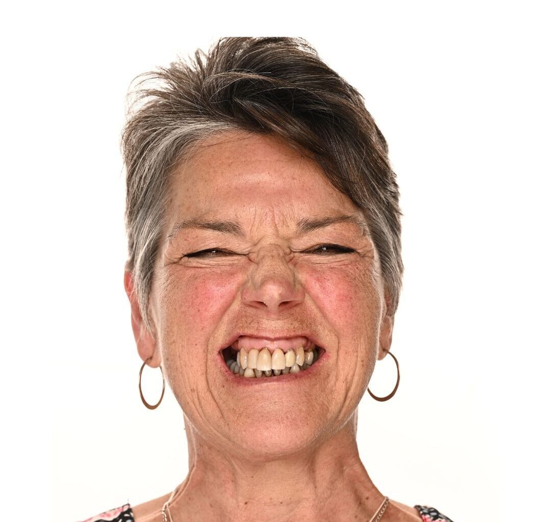 Middle-aged woman with short gray hair and hoop earrings smiling broadly, eyes squinted and nose wrinkled in a joyful, expressive manner against a plain white background.
