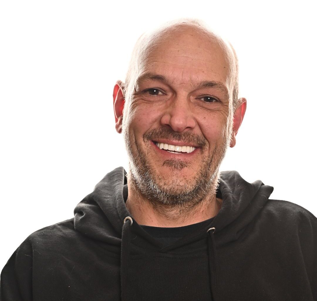 A middle-aged man with a bald head and some facial hair smiles at the camera. He is wearing a black hoodie and is photographed against a plain white background.