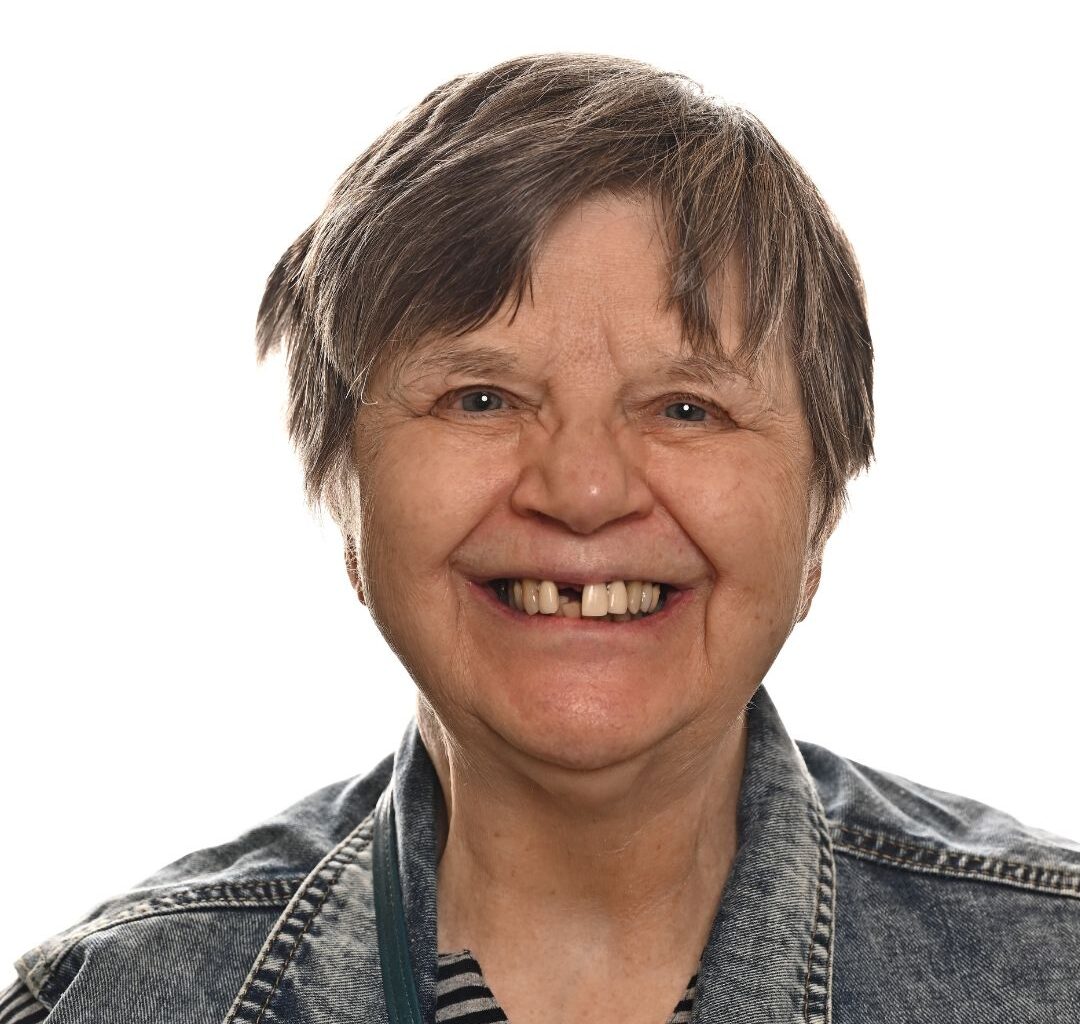 An older woman with short gray hair smiles warmly, showing a gap between her front teeth. She is wearing a denim jacket over a striped shirt, and the background is plain white.