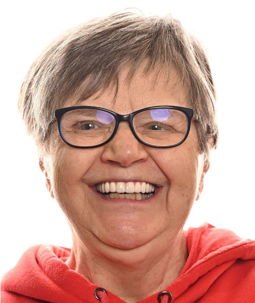 An older woman with short gray hair, wearing black-rimmed glasses and a red hoodie, smiles broadly against a plain white background.