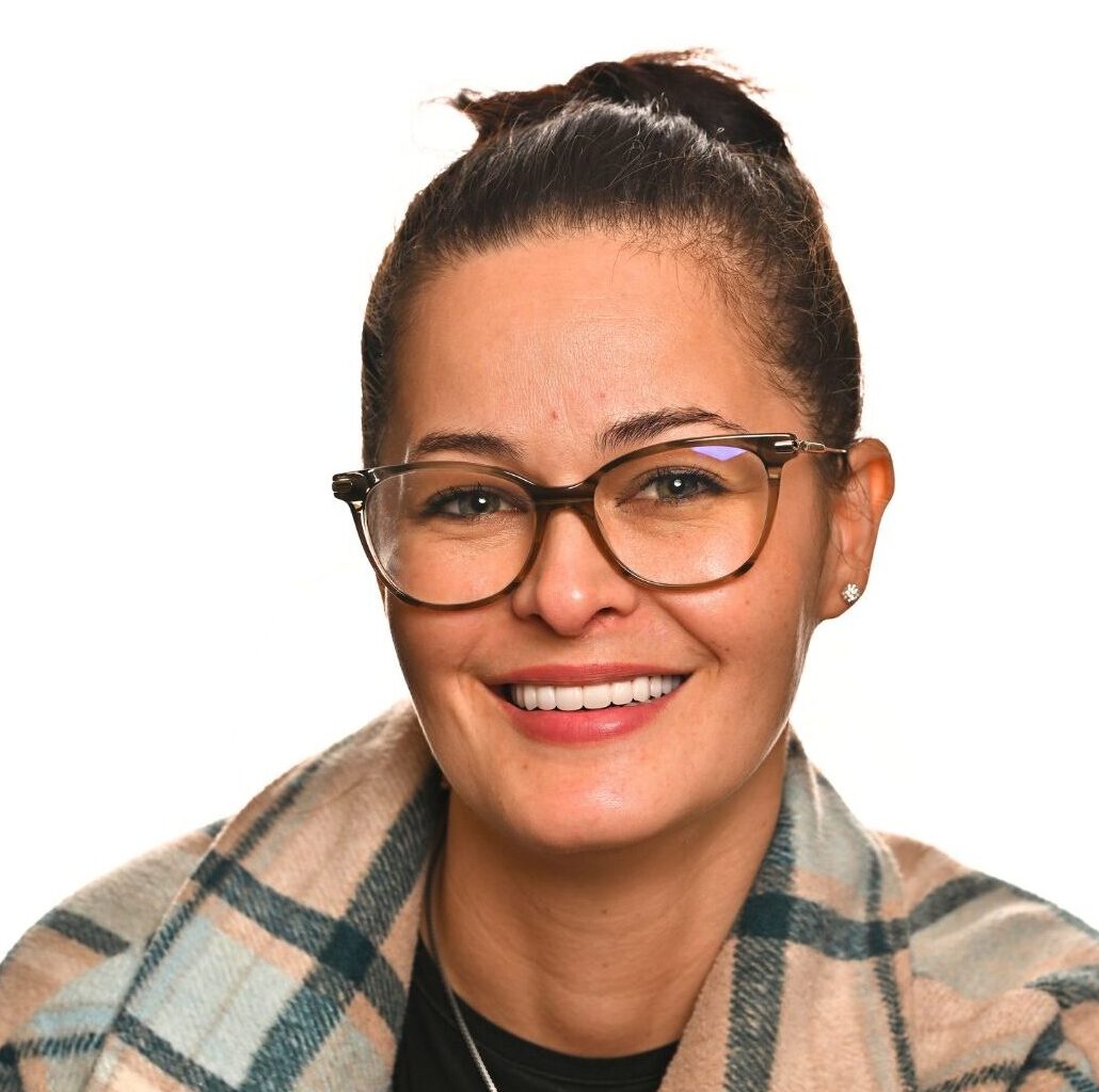 A woman with dark hair in a bun, wearing glasses and a plaid shawl, smiles at the camera against a white background.