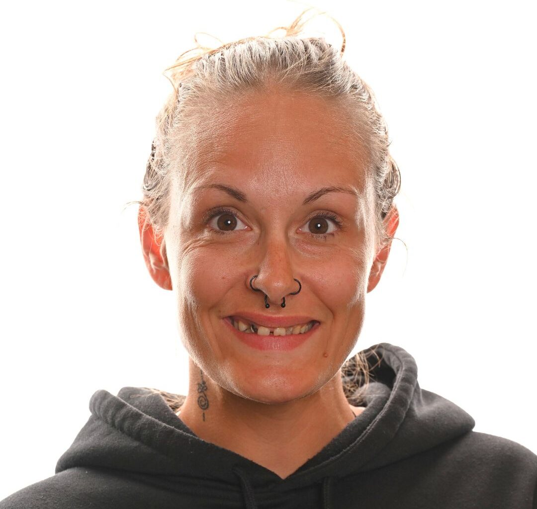 A woman with light skin and pulled-back hair smiles at the camera. She has a nose ring, a missing front tooth, a visible neck tattoo, and is wearing a black hoodie, with a white background behind her.