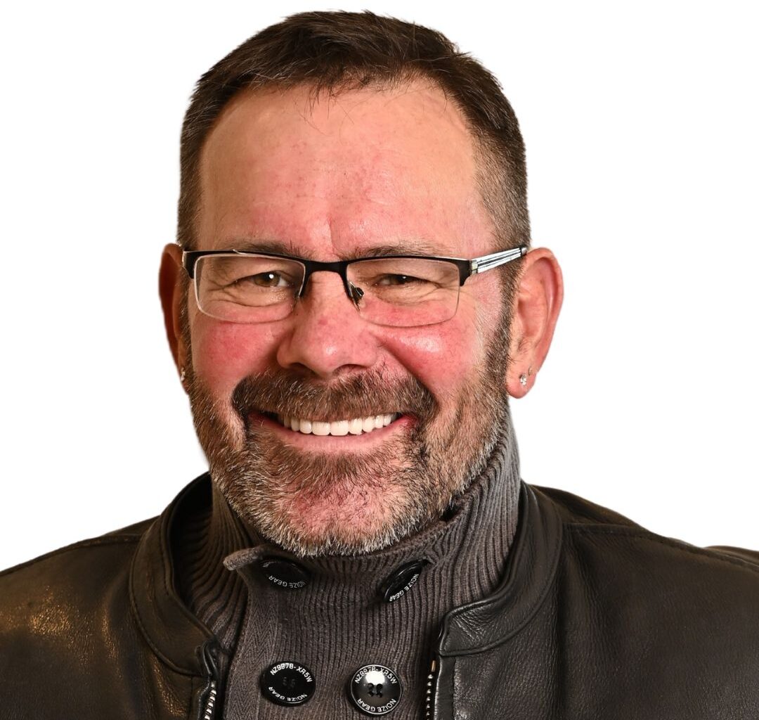 A middle-aged man with short brown hair, glasses, and a beard smiles warmly. He is wearing a gray turtleneck sweater with large buttons and a black leather jacket. The background is plain white.