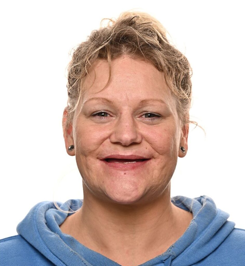 A middle-aged woman with curly blonde hair pulled back, wearing a blue hoodie, smiles at the camera against a plain white background.
