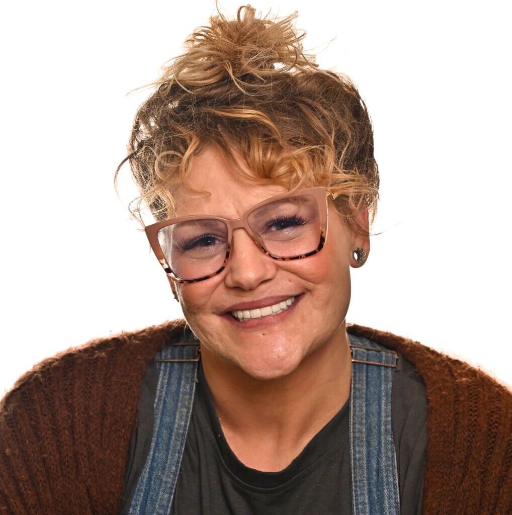 A smiling woman with curly light brown hair in a messy bun, wearing large pink glasses, a brown sweater, denim overalls, and a dark shirt, posing against a white background.