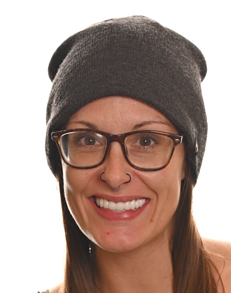 A smiling person with long brown hair, wearing glasses, a dark beanie, and a nose ring, posed against a plain white background.