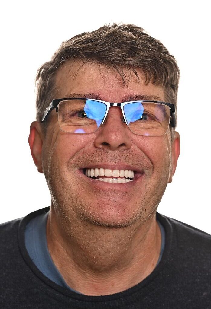 Middle-aged man with short brown hair wearing glasses and a dark sweater smiles broadly at the camera. Reflections are visible on his glasses. The background is plain white.