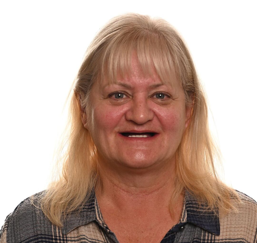 A middle-aged woman with long blonde hair and bangs, wearing a plaid shirt, smiles against a plain white background.