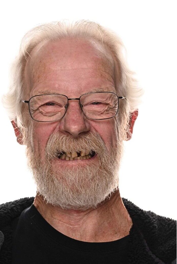 An elderly man with white hair, a full beard, glasses, and uneven teeth smiles at the camera against a plain white background.